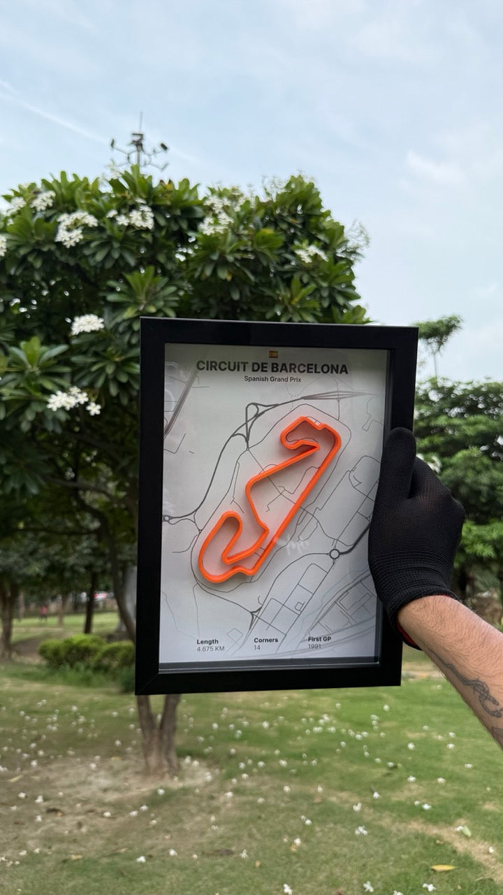 Person holding a framed artwork of Circuit de Barcelona with white background and orange track with trees and grass in the background