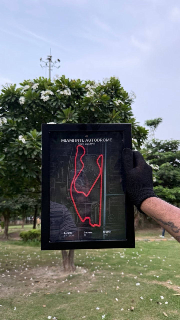 Hand holding a artwork of Miami Intl Autodrome with black background and red track with trees and people in the background