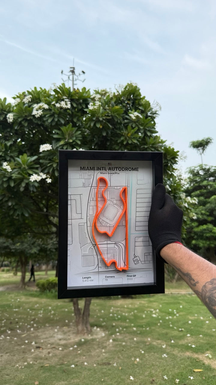 Person holding a framed artwork of Miami Intl Autodrome with white background and orange track with trees and grass in the background