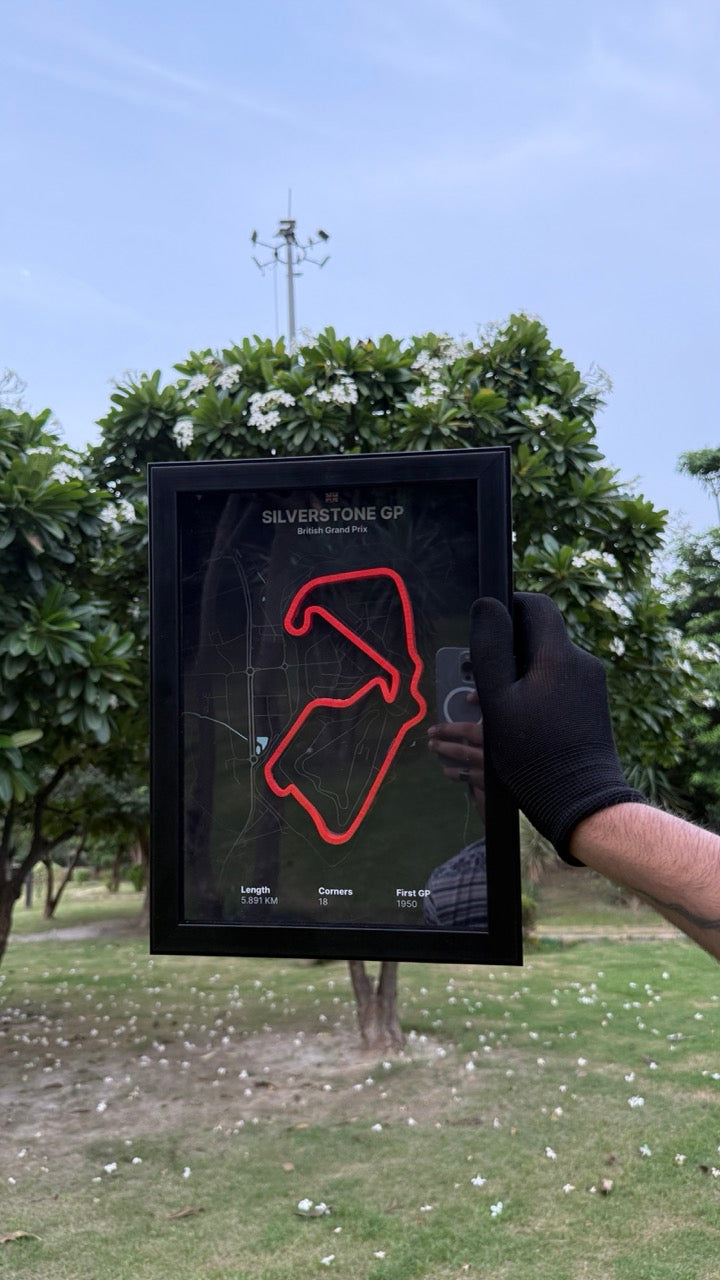 Hand holding a artwork of Silverstone GP with black background and red track with trees and people in the background