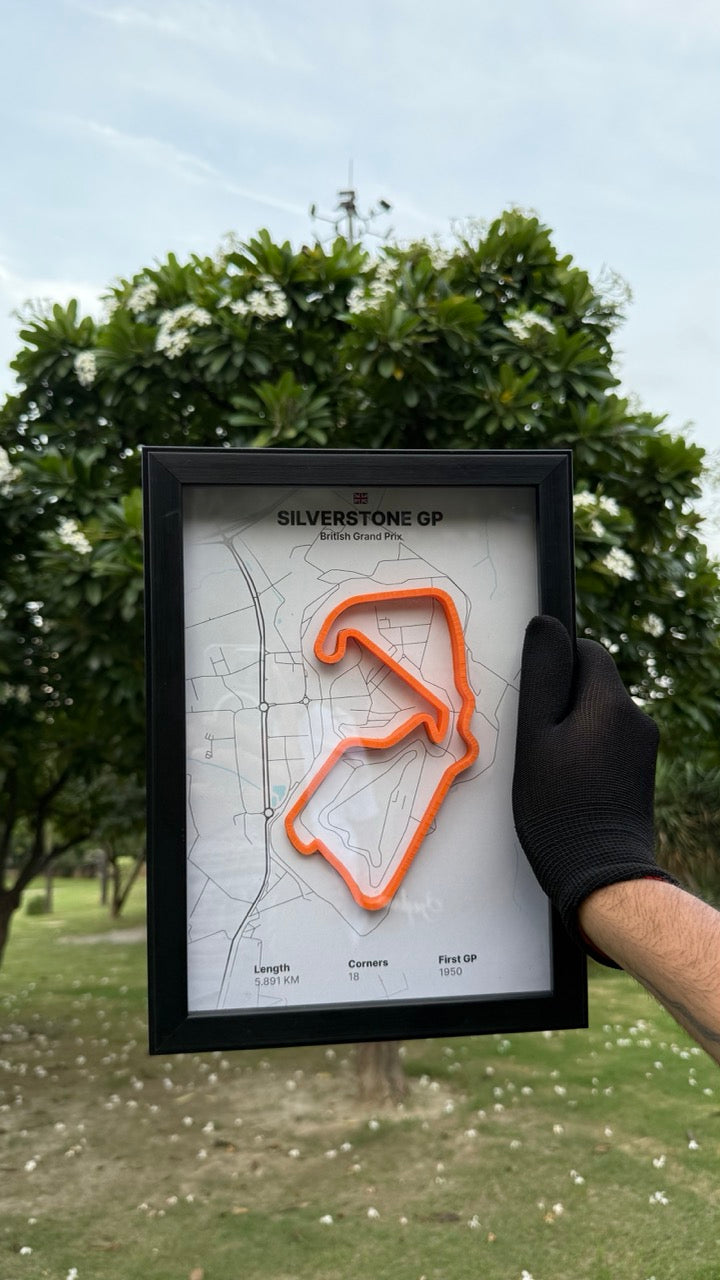 Person holding a framed artwork of Silverstone Circuit with white background and orange track with trees and grass in the background